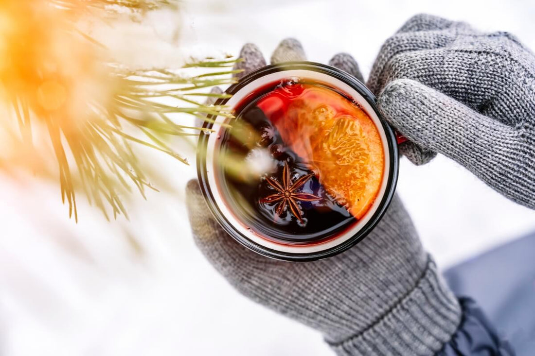 Gloved hands holding a steaming glass of mulled wine with star anise and orange slice in a snowy winter setting